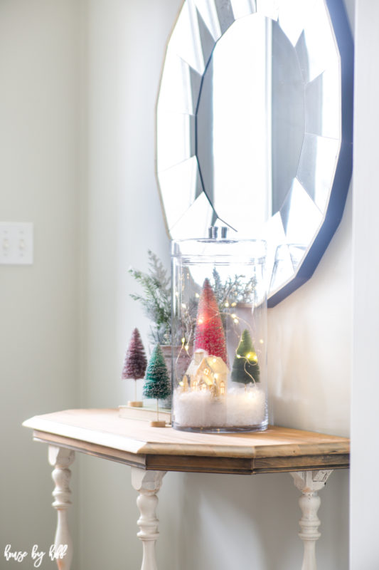 Little trees and a village in a jar on a side table with a mirror above it.