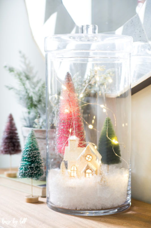 Mini house in a glass jar with trees and lights on the table.