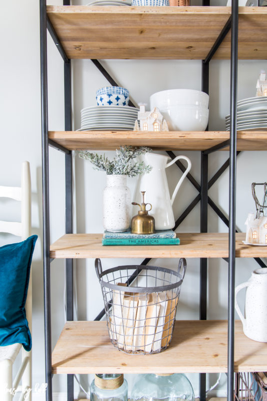 Open shelves with metal basket on the bottom filled with books, a white pitcher on the middle shelf, and plates and bowls on the top shelf.