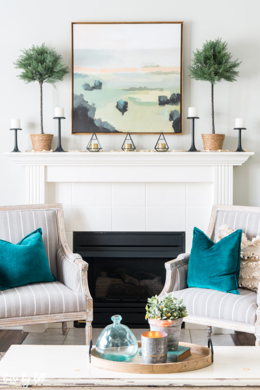 The fireplace mantel with a serene picture above it and topiaries are on either side of the picture.