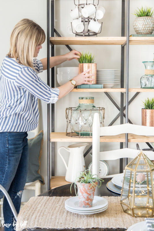 April is fixing a copper vase on the open shelving behind the table.