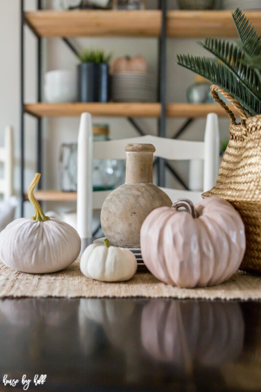 There are three pumpkins, a wooden vase, a stack of plates and a seagrass basket on the table.