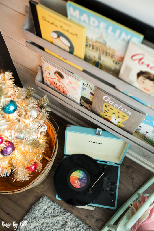 There is a old fashioned record player on the floor beside the books and the tree.